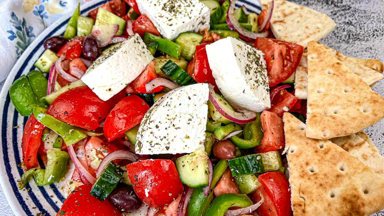 A black bowl contains a Horiatiki salad with blocks of feta cheese, garnished with herbs.