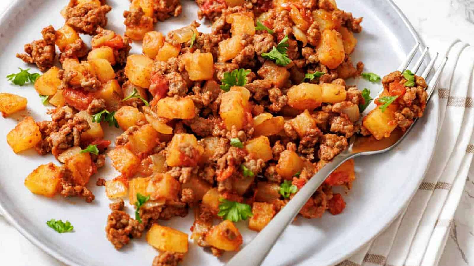 A plate of ground beef and diced potatoes cooked with tomatoes and herbs, garnished with parsley, with a fork on the side.