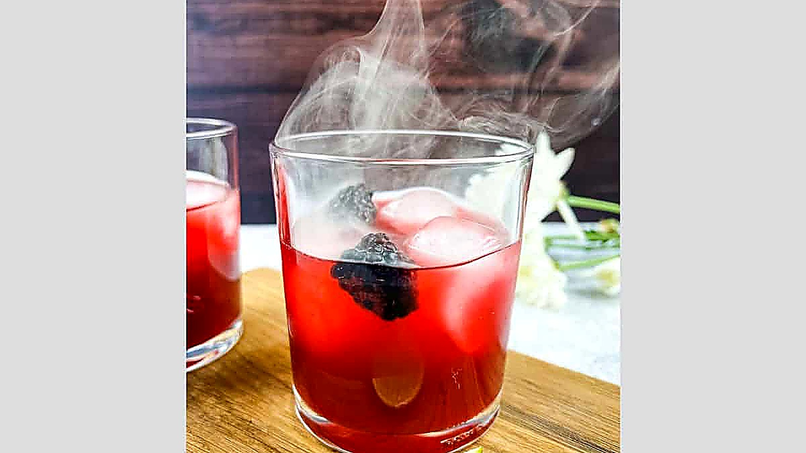 A glass of red drink with ice cubes and blackberries, emitting visible smoke, sits on a wooden surface.