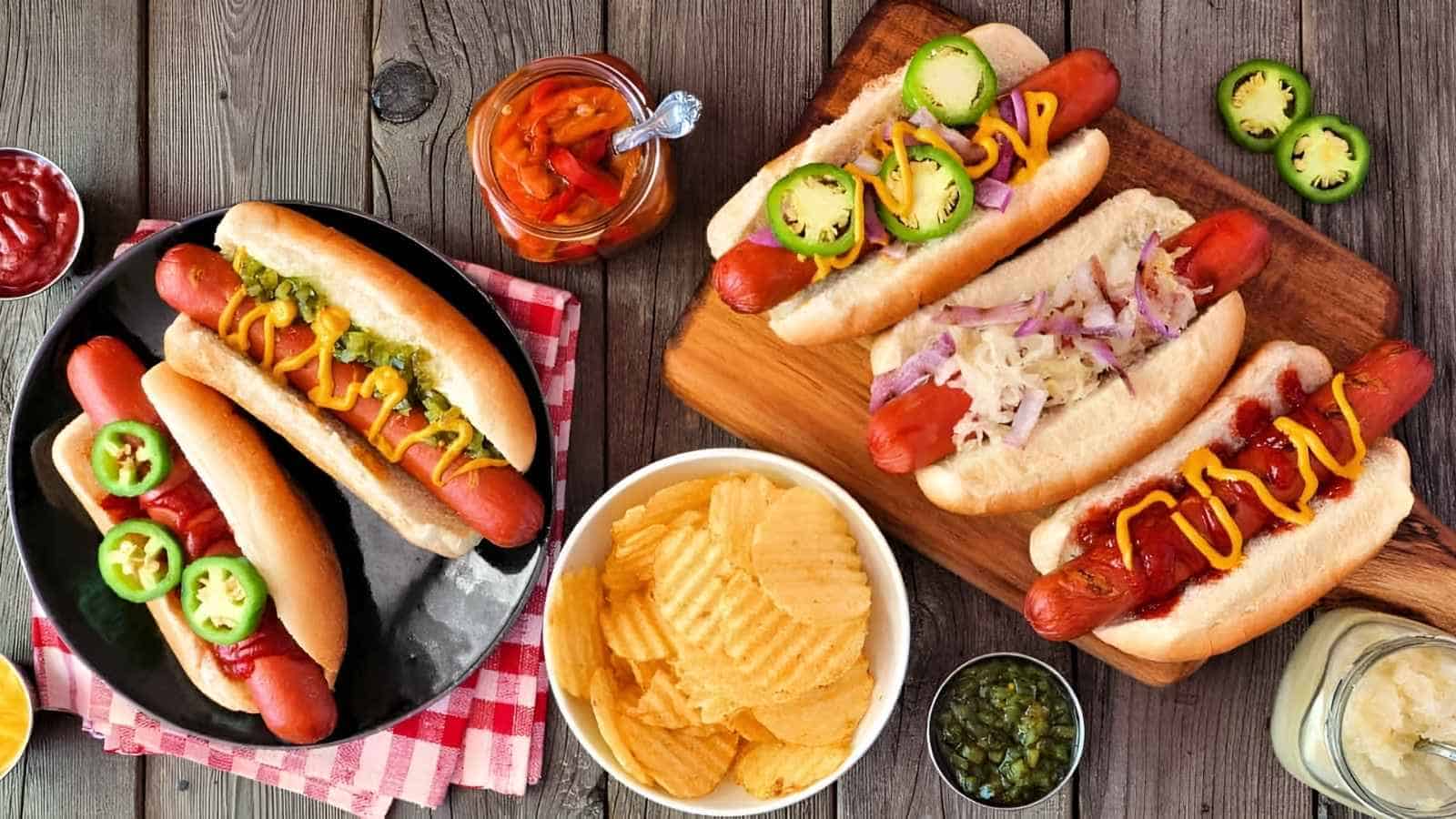 Four hot dogs with various toppings, a bowl of potato chips, sliced jalapeños, relish, and peppers arranged on a wooden table.