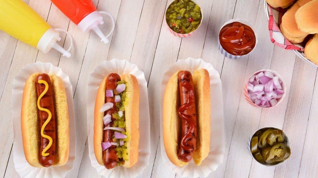 Three hot dogs in buns with various toppings on trays, surrounded by containers of ketchup, mustard, relish, chopped onions, pickles, and a basket of buns on a white wooden surface.