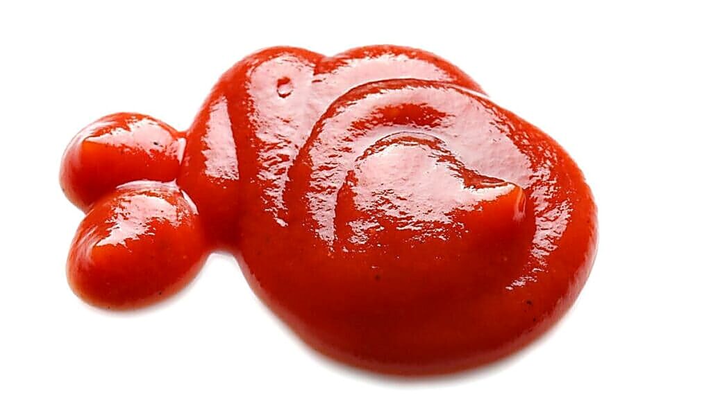 A dollop of ketchup is spread on a white surface, forming an irregular, abstract shape with a glossy texture.