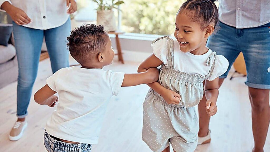 Two young children are smiling and playing together indoors, with two adults standing nearby in a brightly lit room.