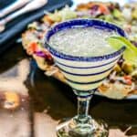 A margarita featuring vibrant margarita flavors, garnished with a lime wedge in a blue-rimmed glass, sits on a table next to salt and pepper shakers and a plate of nachos topped with vegetables.