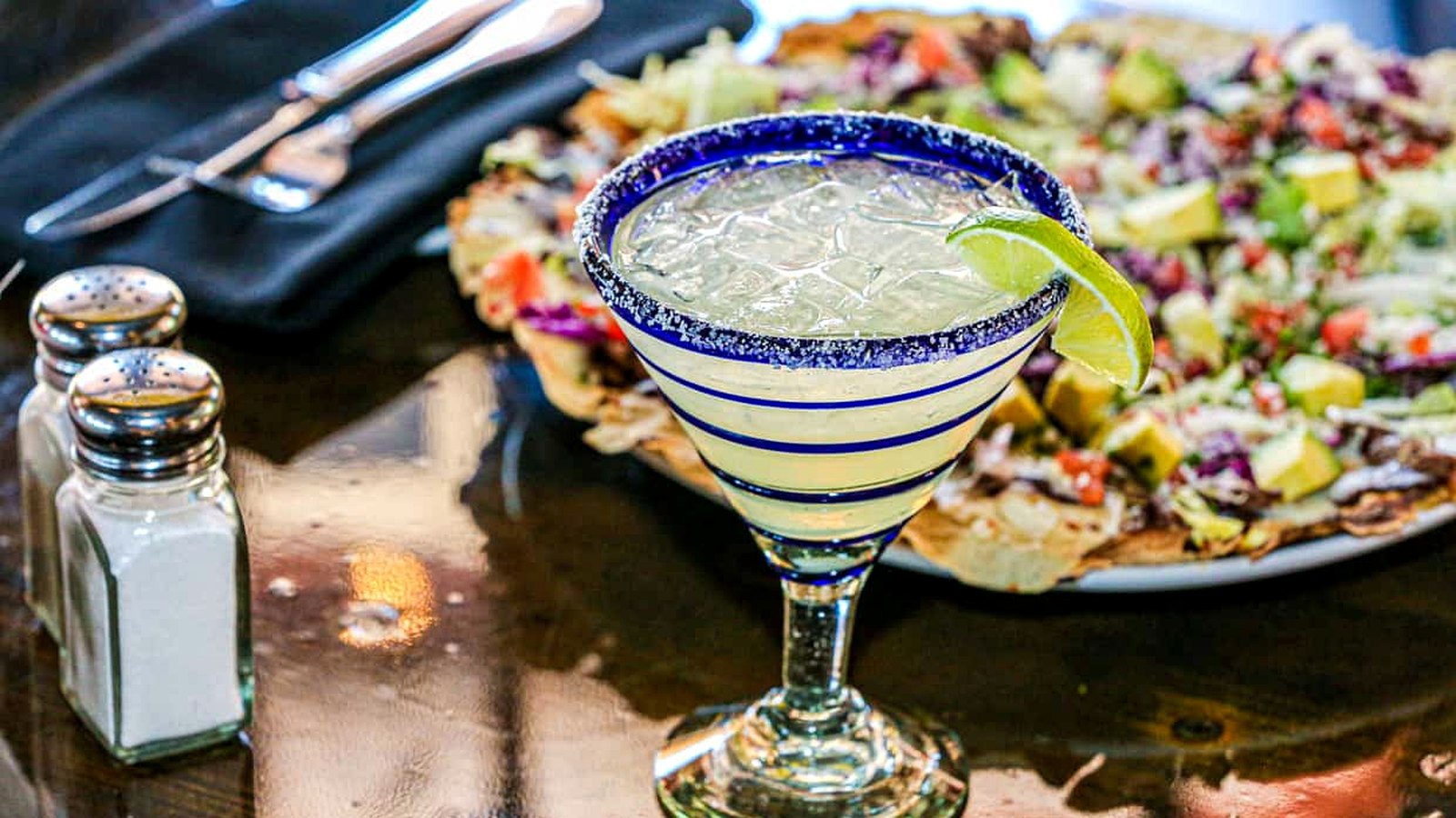 A margarita featuring vibrant margarita flavors, garnished with a lime wedge in a blue-rimmed glass, sits on a table next to salt and pepper shakers and a plate of nachos topped with vegetables.