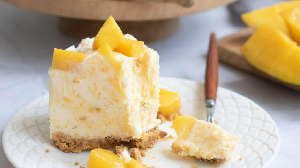 A square slice of mango cheesecake with a crumbly crust, topped with mango pieces, sits on a white plate beside a fork.
