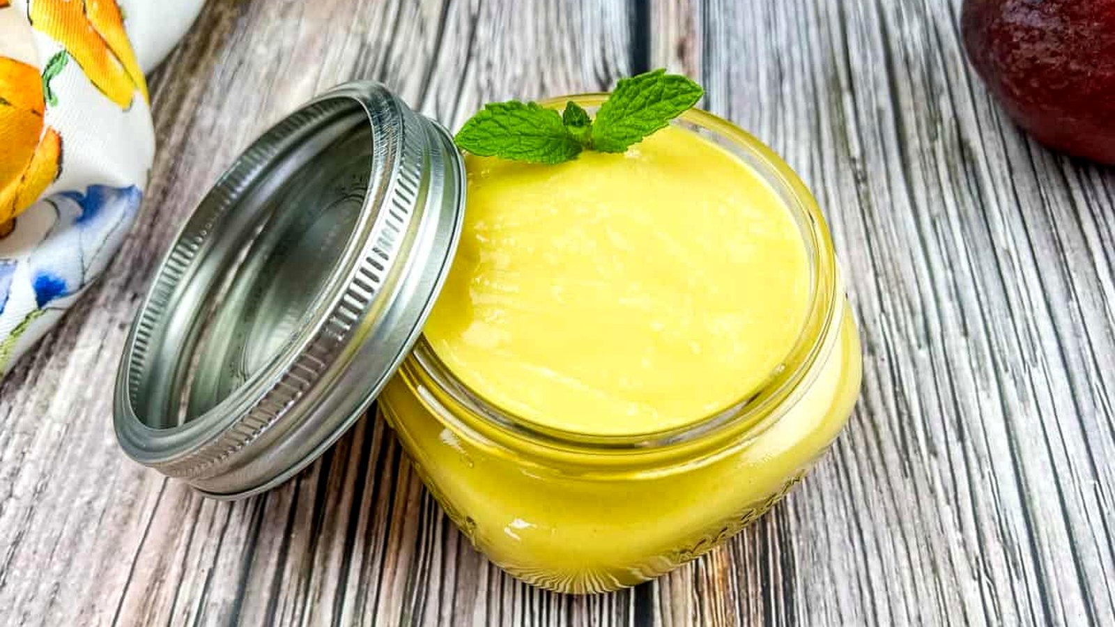 A jar of mango curd next to a mango and a mint leaf.