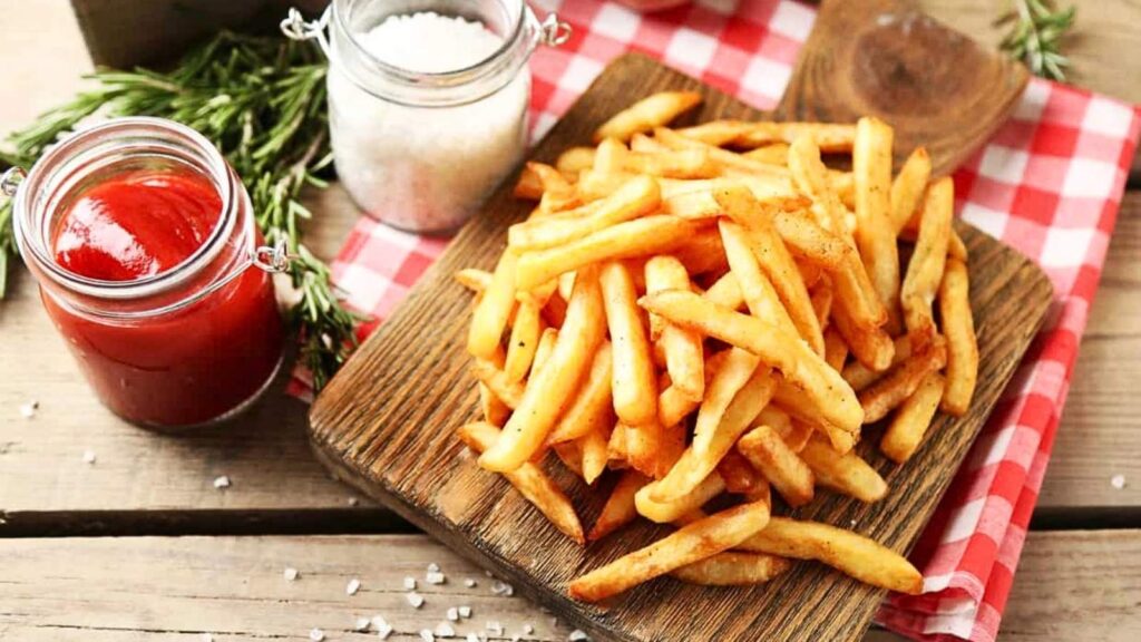 French fries on a wooden board with a jar of ketchup, salt, and rosemary rest on a red and white checkered cloth&mdash;perfect for celebrating National French Fry Day.