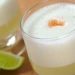 A close-up of two glasses of frothy, pale yellow cocktail with a drop of bitters on top, accompanied by lime wedges on a wooden surface.