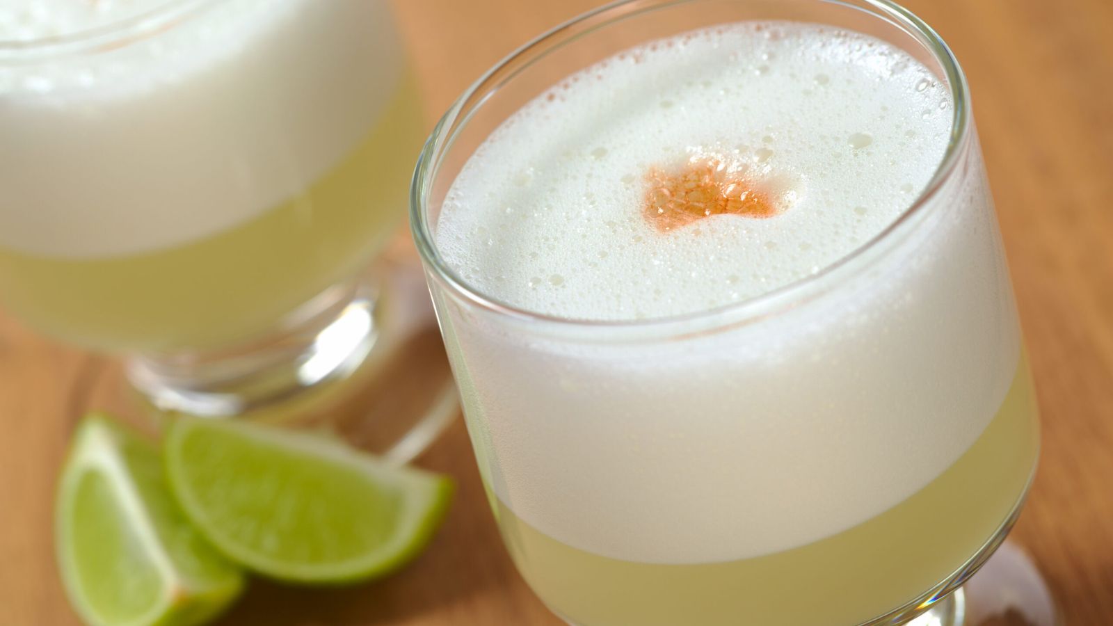 A close-up of two glasses of frothy, pale yellow cocktail with a drop of bitters on top, accompanied by lime wedges on a wooden surface.