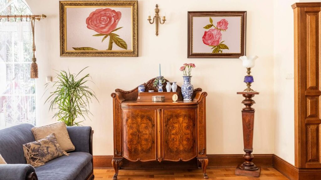 A living room with vintage wooden furniture, floral paintings on the wall, decorative vases, and a blue sofa with cushions.