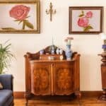 A living room with vintage wooden furniture, floral paintings on the wall, decorative vases, and a blue sofa with cushions.