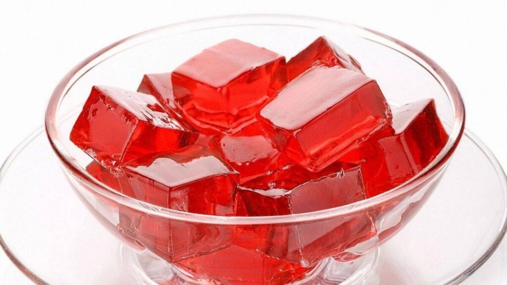 A clear glass bowl filled with red gelatin cubes is placed on a matching glass saucer against a white background.