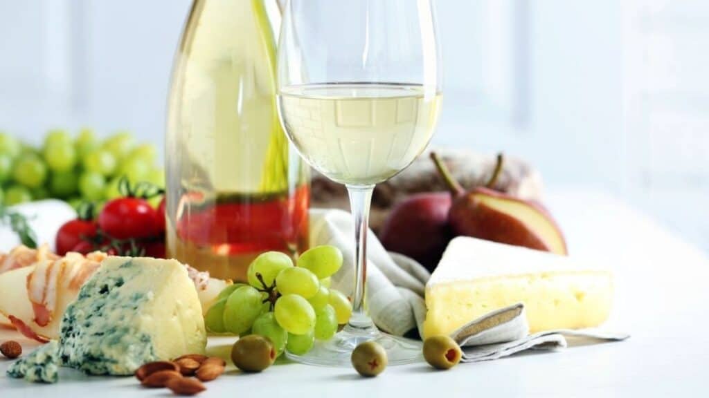 A glass of white wine, cheese, green grapes, cherry tomatoes, almonds, olives, a pear, and a bottle of wine arranged on a white table.