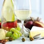 A glass of white wine, cheese, green grapes, cherry tomatoes, almonds, olives, a pear, and a bottle of wine arranged on a white table.