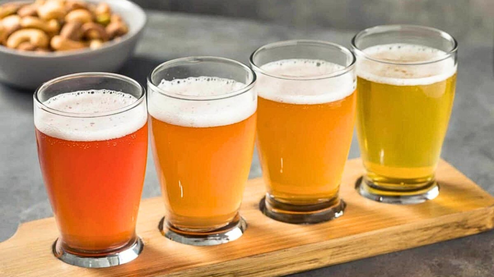 Four glasses of different colored beers are arranged in a row on a wooden tray, with a bowl of mixed nuts in the background&mdash;a perfect setting to compare tasting notes or even discuss tequila prices with friends.
