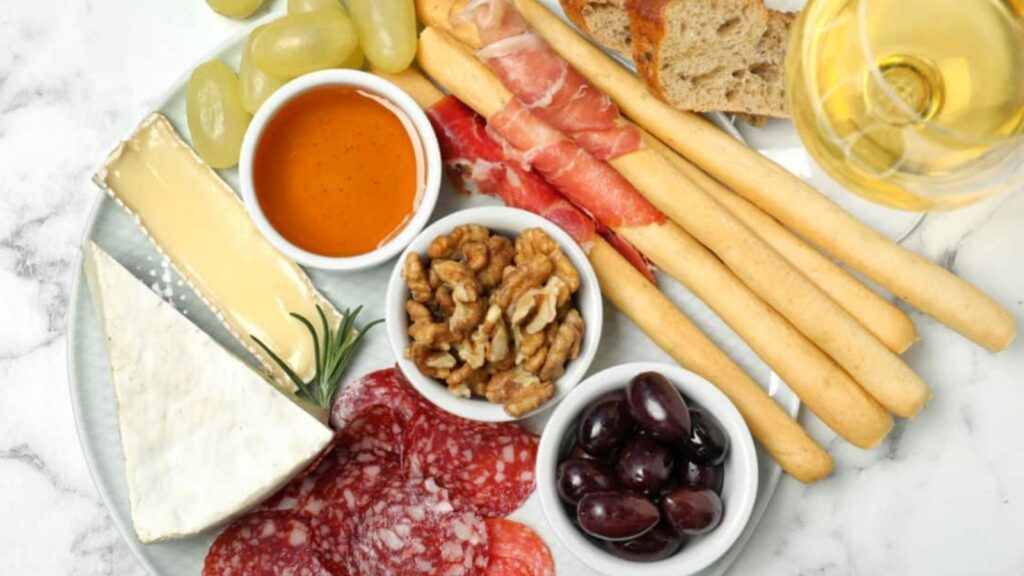 Charcuterie board with brie, salami, prosciutto, grapes, walnuts, olives, breadsticks, honey, bread, and a glass of white wine on a marble surface.