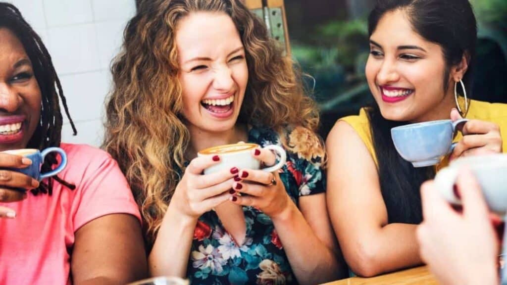 Three women sit together at a table, smiling and holding coffee cups, enjoying a casual conversation in a bright setting.