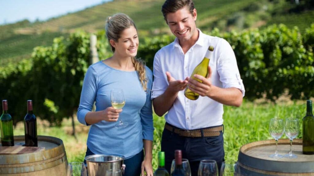A man and woman stand outdoors by wine barrels in a vineyard, holding glasses and examining a wine bottle.