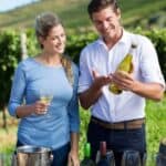 A man and woman stand outdoors by wine barrels in a vineyard, holding glasses and examining a wine bottle.