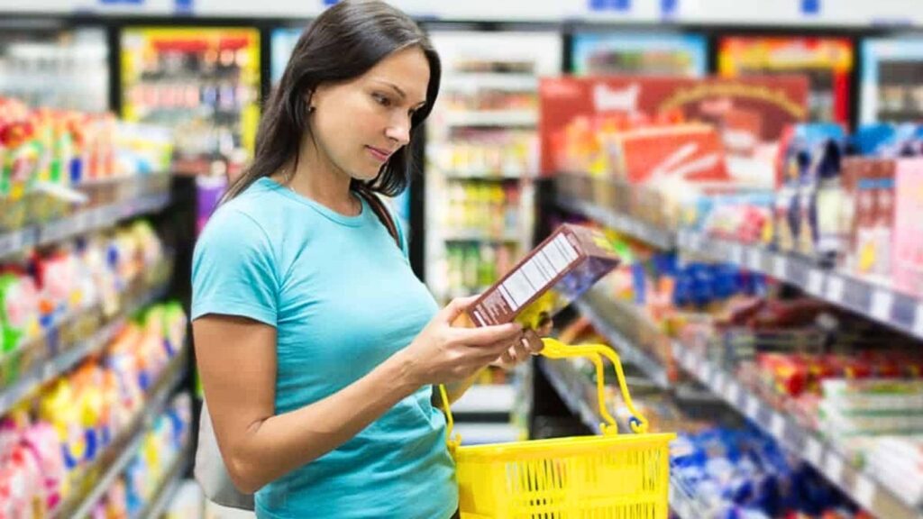 Woman in a blue shirt holds a yellow shopping basket and examines a packaged product in a grocery store aisle, carefully checking the label for compliance with the new California food law.