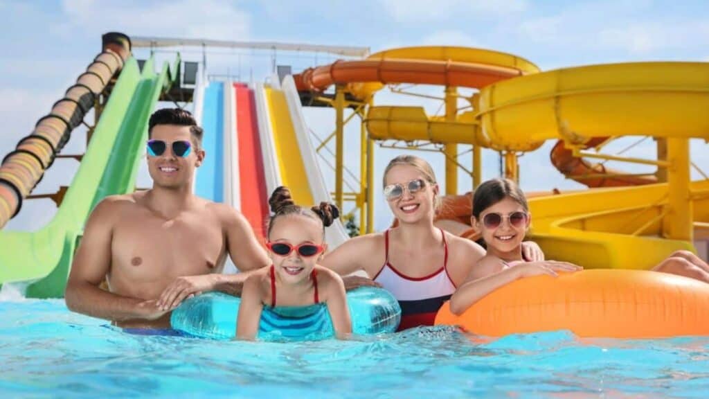 A family of four wearing swimsuits and sunglasses smiles in a pool, with water slides and colorful tubes in the background on a sunny day.