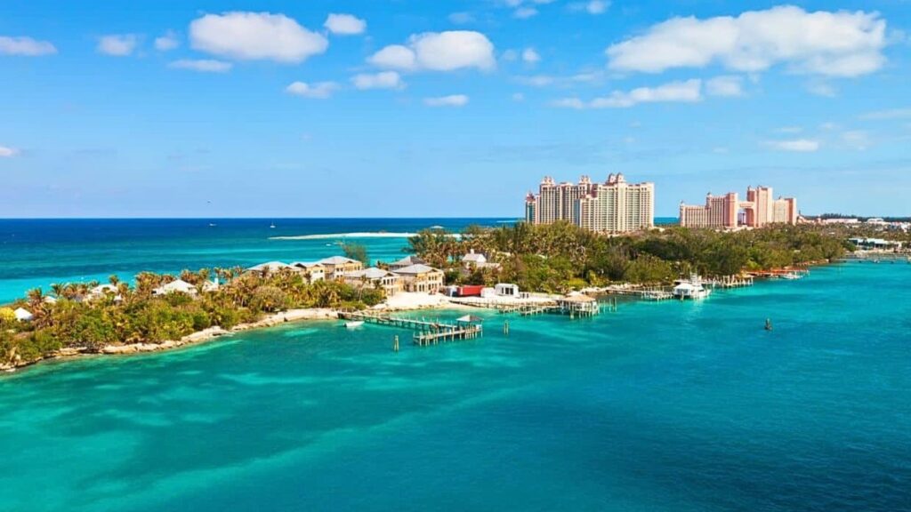 Aerial view of a coastal resort area with tall buildings, lush greenery, small docks, and turquoise ocean water under a partly cloudy sky, reminiscent of the rich history of Nassau.