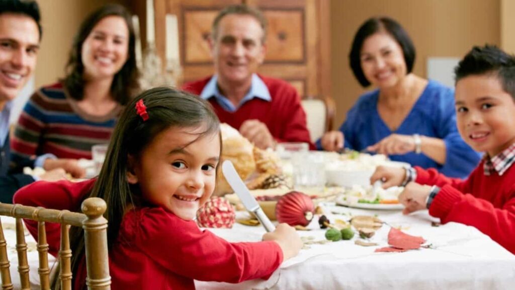 Six people of different ages sit at a festive dining table, smiling and eating together. The group includes a young girl in the foreground holding a knife and facing the camera.