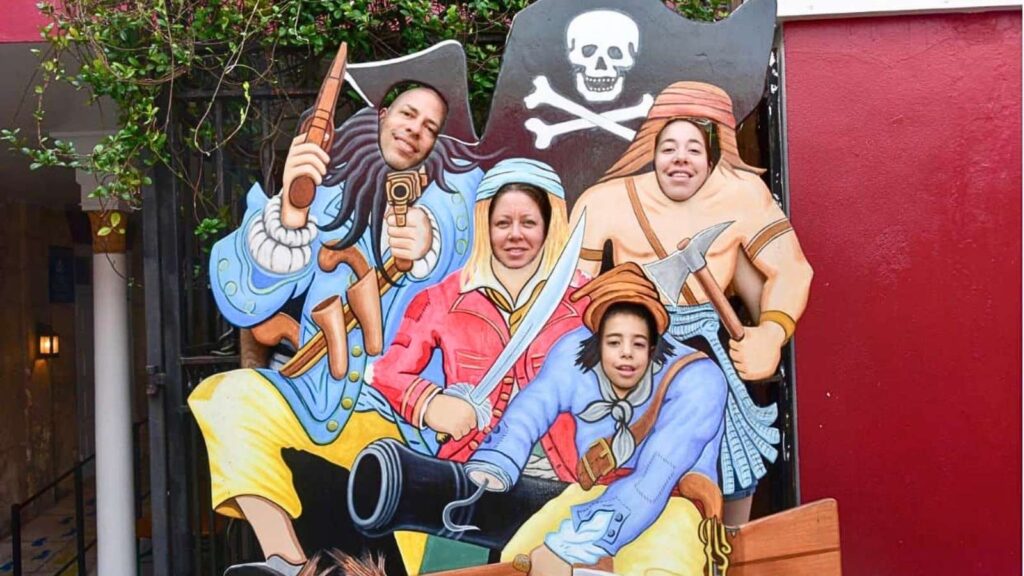 Four people pose with their faces in a pirate-themed cutout board featuring colorful pirate costumes and props, including swords and a cannon&mdash;evoking the swashbuckling history of Nassau and its legendary pirates.