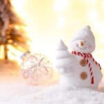 A white snowman figurine with a red striped scarf holds a tree, surrounded by snow, a glass ornament, and blurred lit trees in the background.