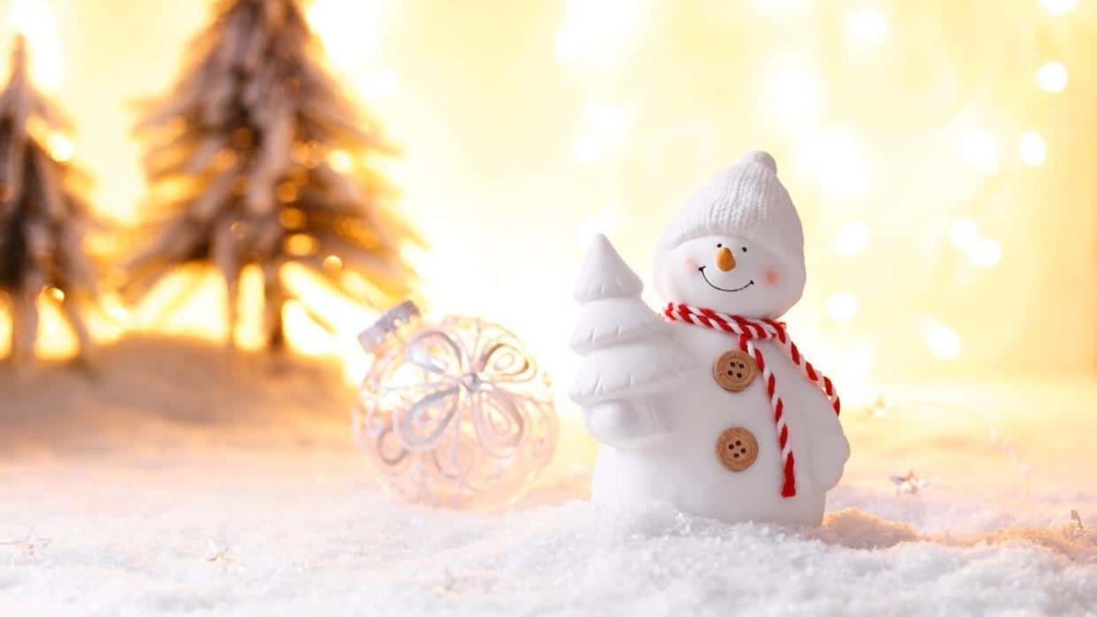 A white snowman figurine with a red striped scarf holds a tree, surrounded by snow, a glass ornament, and blurred lit trees in the background.