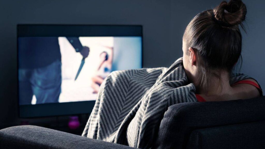A person wrapped in a blanket sits on a couch watching a suspenseful scene with a knife on TV.