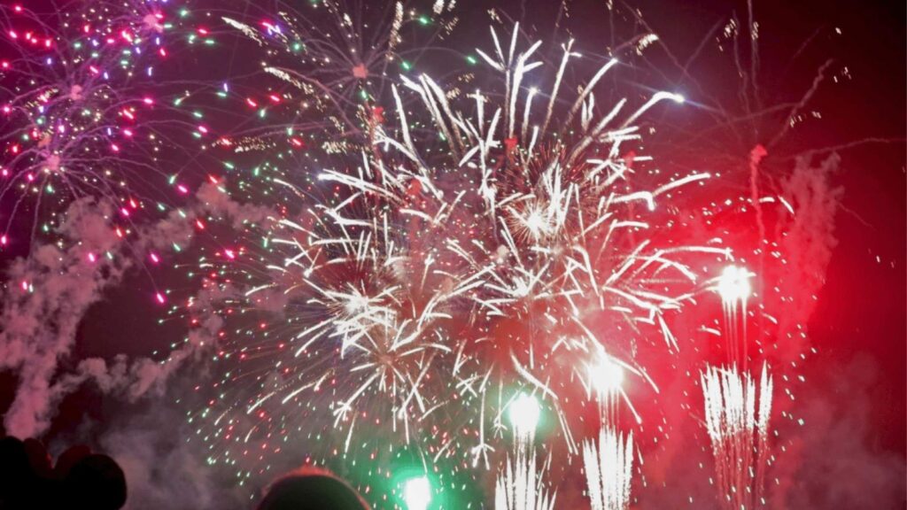 Fireworks burst in the night sky with red, green, and white colors. Silhouetted heads of spectators are visible at the bottom of the image.