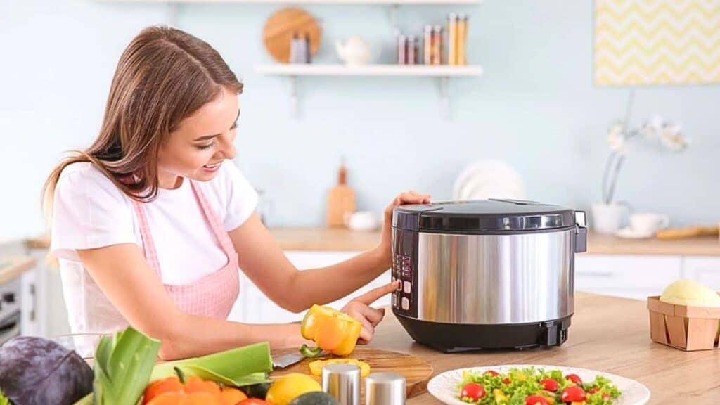 A woman in a kitchen presses a button on an electric rice cooker, with various vegetables and a prepared salad on the counter.
