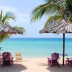 Four purple chairs and two thatched umbrellas on a sandy beach face clear blue water, with palm trees and lounge chairs in the background under a bright sky—an inviting scene perfect for enjoying exclusive Marriott summer deals.