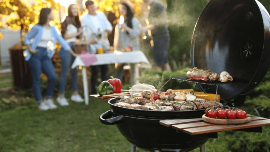 People stand and talk near a table in the background while food cooks on a grill in the foreground in a backyard setting, where everyone enjoys friendly conversation and follows good cookout etiquette.