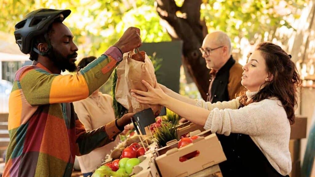 A man in a helmet receives a paper bag from a woman at an outdoor market stand with fresh produce; another man stands in the background.