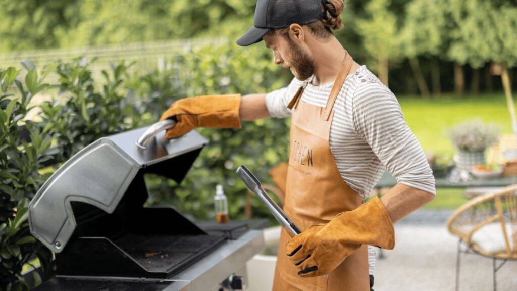 A man wearing an apron and gloves opens a grill outdoors while holding a grill brush, with greenery in the background, getting ready to savor the moment despite rising cookout costs.