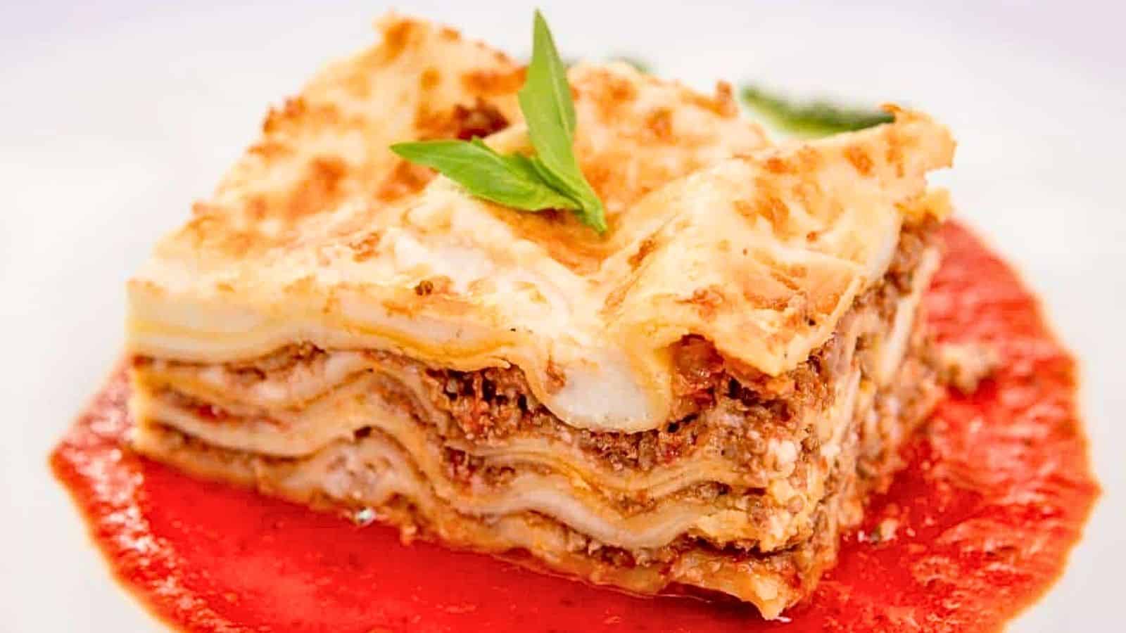 A slice of lasagna with visible meat and cheese layers sits on a pool of red tomato sauce, garnished with a small basil leaf&mdash;a perfect comfort dish to celebrate National Lasagna Day.
