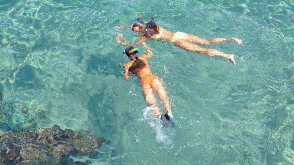 Two people wearing snorkels swim next to each other in clear, turquoise water near some rocks.