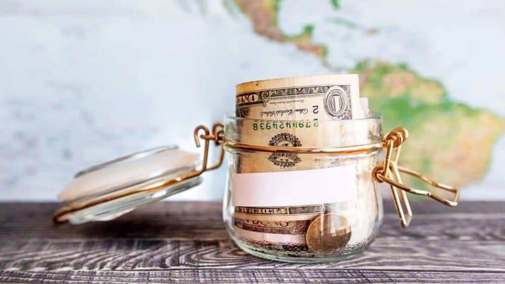 A glass jar filled with U.S. dollar bills and coins sits on a wooden surface, with a blurred map in the background&mdash;an emblem of saving for boomcession travel plans ahead.