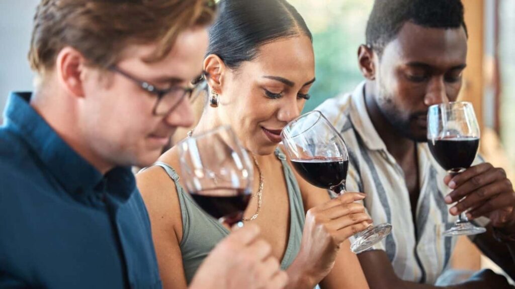 Three people sit side by side, holding glasses of red wine, each smelling the wine with focused expressions.