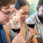Three people sit side by side, holding glasses of red wine, each smelling the wine with focused expressions.