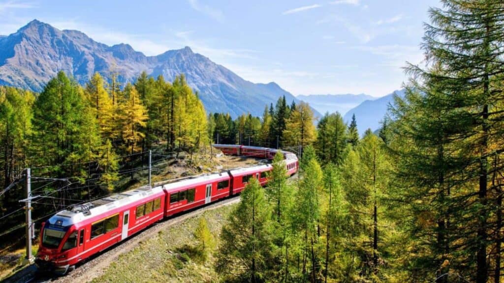 A red passenger train travels through a forested mountain landscape with tall trees and distant peaks under a clear blue sky.