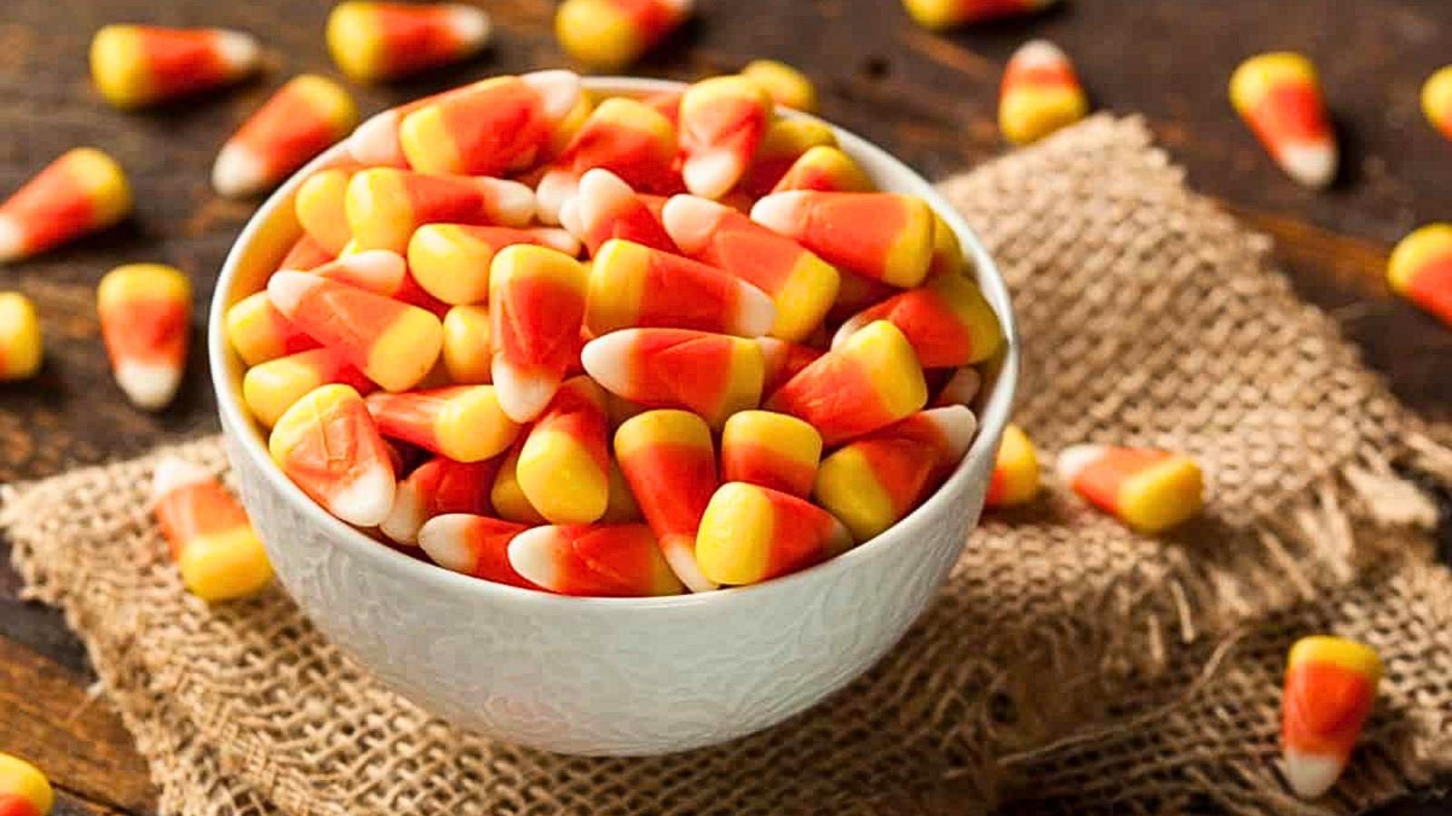 A white bowl filled with candy corn sits on a piece of burlap, with additional candy corn scattered on the wooden surface nearby.