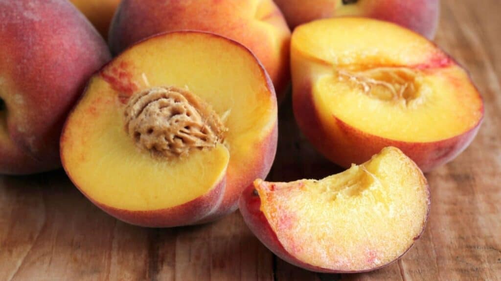 Several ripe peaches on a wooden surface, with one peach cut in half showing the pit and a segment removed.