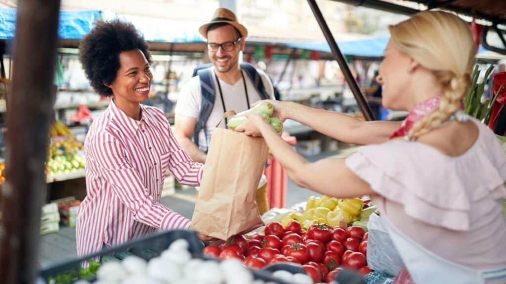 A vendor hands vegetables in a paper bag to a smiling customer at an outdoor market, with another person standing nearby.