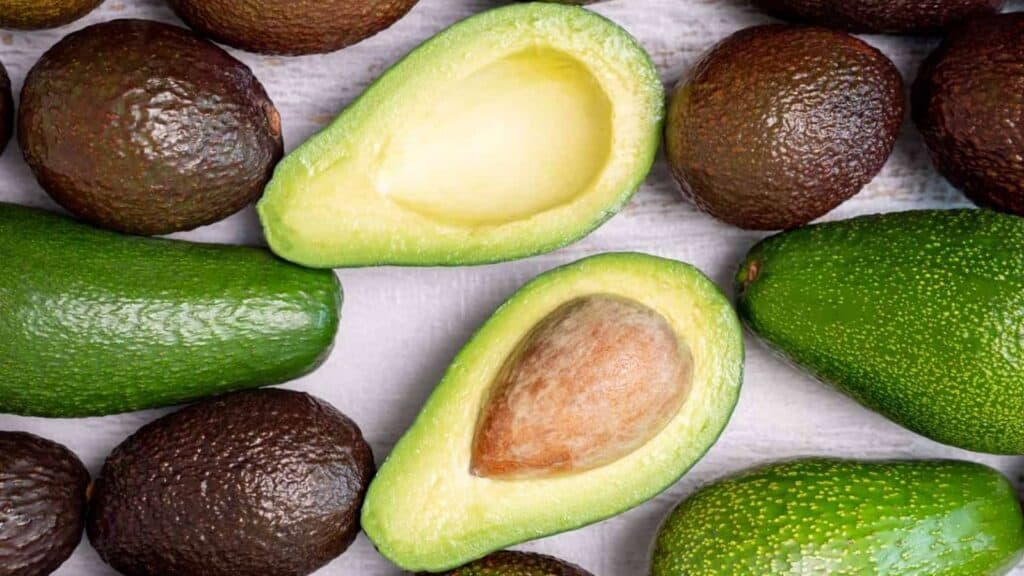 Several whole avocados and one avocado cut in half, showing the seed and green flesh, are arranged on a light surface—showcasing the perfect results achieved by an advanced avocado harvesting robot.