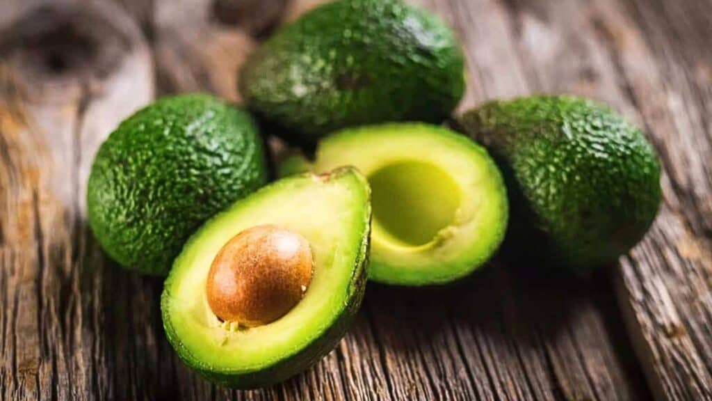 Four avocados on a wooden surface, with one cut in half showing the seed and green flesh—an inviting arrangement that highlights the perfect produce often selected by an avocado harvesting robot.
