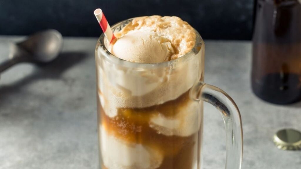 A glass mug filled with root beer and scoops of vanilla ice cream, topped with foam and a red-striped straw on a gray surface.